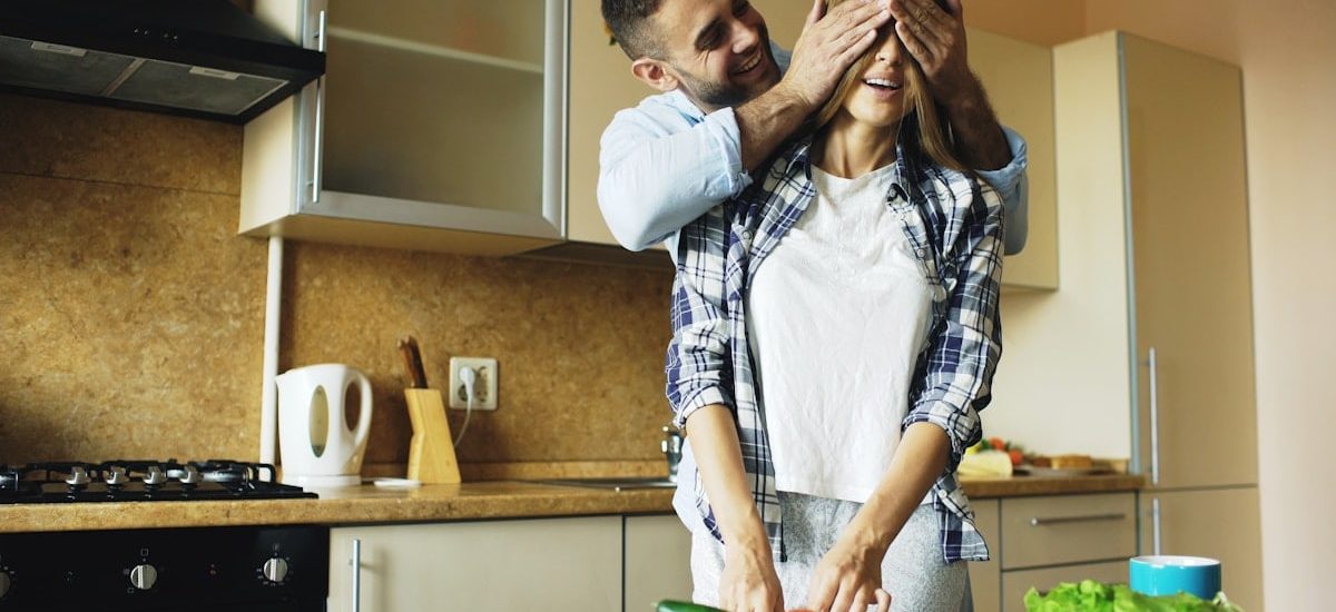 Young man covering girlfriends eyes with hands and surprising her in the kitchen at home. Loving couple cooking breakfast in the morning