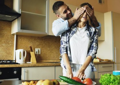 Young man covering girlfriends eyes with hands and surprising her in the kitchen at home. Loving couple cooking breakfast in the morning